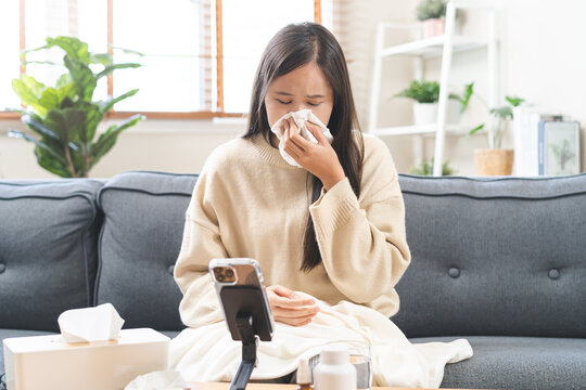 Sick young Asian woman holding thermometer and using tissue sneezing during online video call medical consultation with doctor on smartphone at home, concept of telemedicine healthcare technology