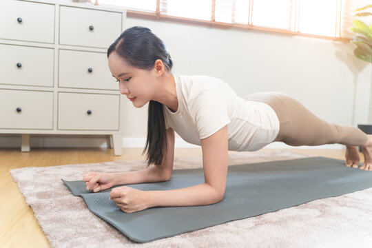 Determined asian young woman practicing forearm plank exercise on mat at home living room for core strength training fitness workout and healthy wellness lifestyle building
