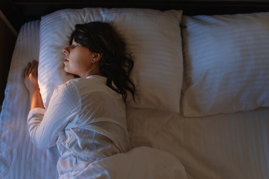 Deep sleep, sleep phases, recovery. A woman with dark curly hair is sleeping in bed, wearing light-colored pajamas, on white striped bedding.