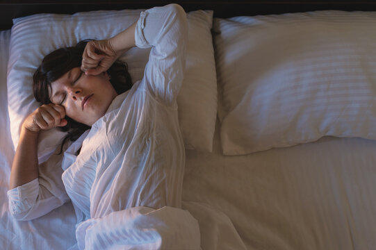 A woman with dark curly hair is sleeping in bed, wearing light-colored pajamas, on white striped bedding. She Rubs their eyes with their hands and wakes up.