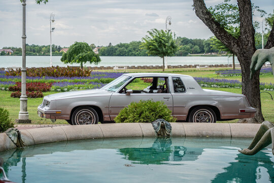 Side view profile of silver 1981-1986 Oldsmobile Cutlass Supreme Brougham 4th gen classic coupe behind park fountain pool, Palic, Serbia, Jul 5, 2026