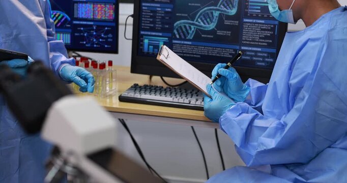 Medical researchers in protective clothing review genetic data and laboratory notes beside a microscope in a modern science lab.
