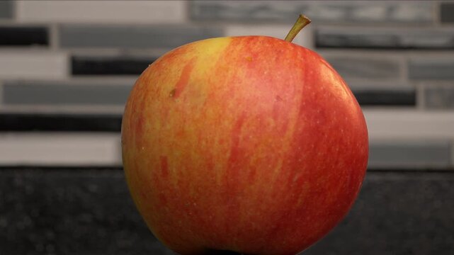 360 degree view of an imperfect gala apple on a black counter with tiled backdrop. 