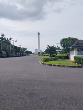 Scenic view of the National Monument (Monas) in Jakarta, Indonesia, with an asphalt road, military trucks, and green trees.
