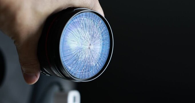 Close-up of a hand holding a camera lens with a cracked front element against a dark background, damage, repair needs, and fragile equipment.