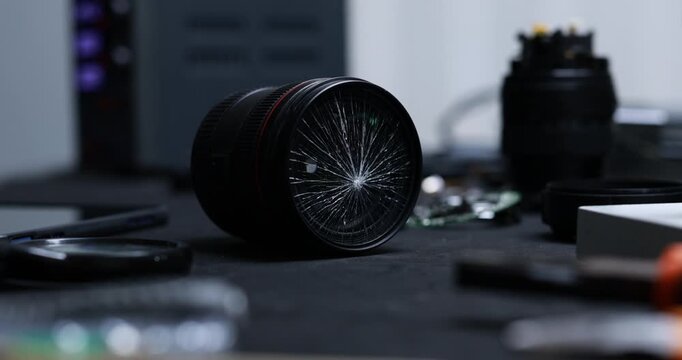 Close-up of a cracked camera lens lying on a dark workbench with blurred photography gear and tools in the background.