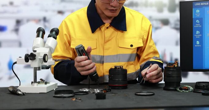 Technician in yellow safety uniform repairs a camera lens at a workbench using a microscope, precision tools and spare parts beside a repair screen.