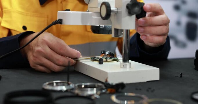 Watchmaker in a yellow work coat repairs a camera lens under a precision magnifier, with small tools and lens parts on a dark workbench.