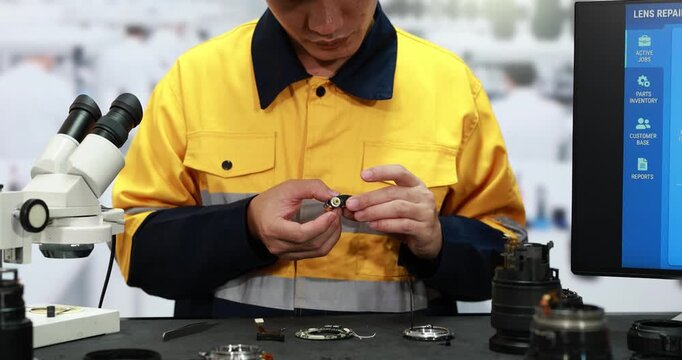Technician in yellow work uniform repairing a small camera part at a bench with microscope, lenses and computer screen in a repair workshop