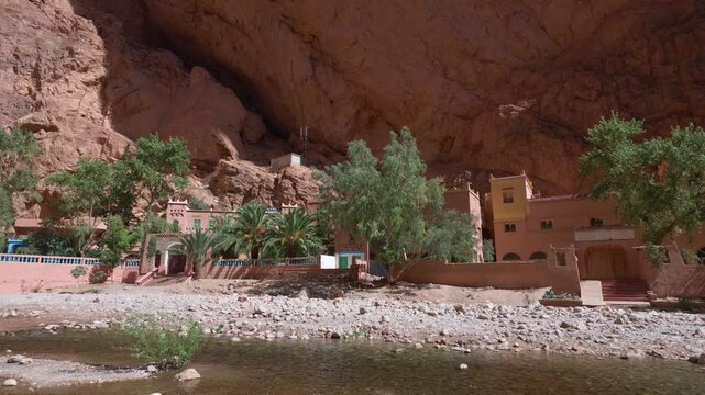 Traditional moroccan kasbah nested at the base of the todra gorge canyon with a flowing river in the foreground, showing the scenic landscape of the atlas mountains