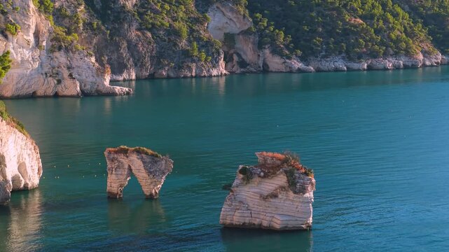 Baia delle Zagare iconic Faraglioni rock stacks aerial 70mm view in Gargano. Cinematic drone slow flyby over pink sunrise morning light empty cove 4K