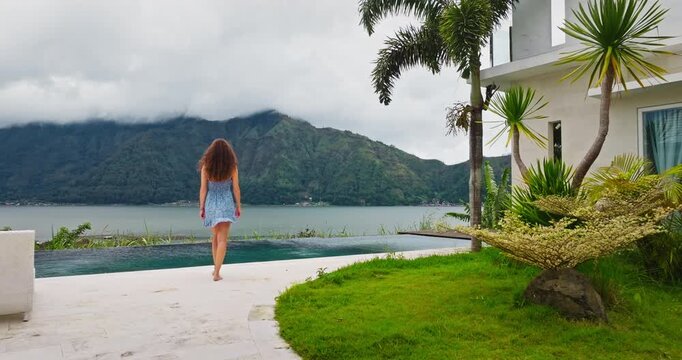 Young woman in blue dress barefoot walking modern resort to infinity pool edge, enjoy view on vast Batur Lake and green mountains, experiencing travel, relaxation and natural beauty of Bali island