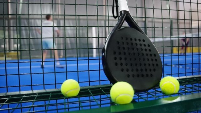Сlose-up shot focused on a professional padel tenis racket and neon yellow balls resting in the foreground and paddle players are on blurred background. 