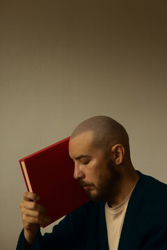 Side view portrait of a bald man with beard resting his head against a red vintage hardcover book with closed eyes, conceptual shot of peaceful reading, education and storytelling, studio background.