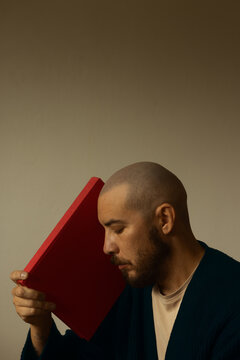 Side view portrait of a bald man with beard holding a red hardcover book against his head with closed eyes, peaceful quiet reading concept, literary education background on neutral studio wall.