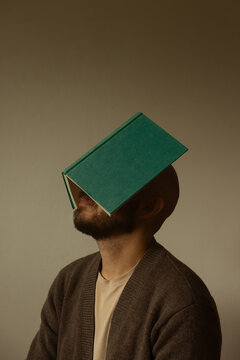 Three-quarter portrait of a bald man with beard balancing an open green hardcover book on his face, conceptual shot of reading fatigue, knowledge overload and literature, beige studio background.