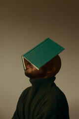 Side profile portrait of a bald man with beard balancing an open green hardcover book over his face, conceptual shot of knowledge overload, reading and literature, studio beige background. © Francis