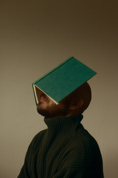 Side profile portrait of a bald man with beard balancing an open green hardcover book over his face, conceptual shot of knowledge overload, reading and literature, studio beige background.
