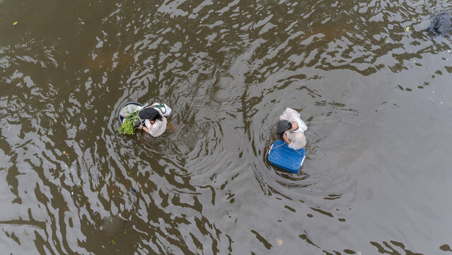 Aerial view of two people wading through deep floodwaters carrying containers and fresh produce