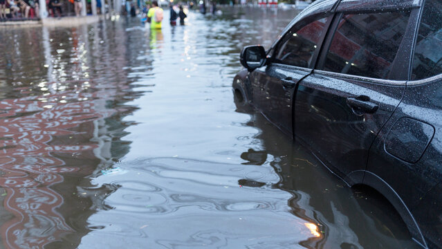 A dark car parked on a flooded city street during a heavy rainstorm with people in the distance