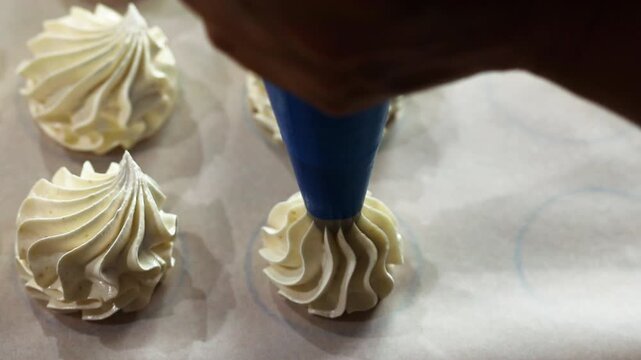 Baker's hand with blue piping bag creates decorative meringue peaks on parchment paper, showcasing the intricate design process in a well-lit kitchen environment