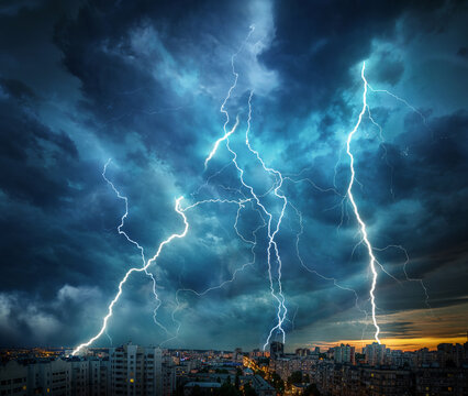 Dramatic evening cityscape illuminated by numerous bright lightning strikes across a dark stormy sky, with houses and urban buildings glowing with warm city lights. 