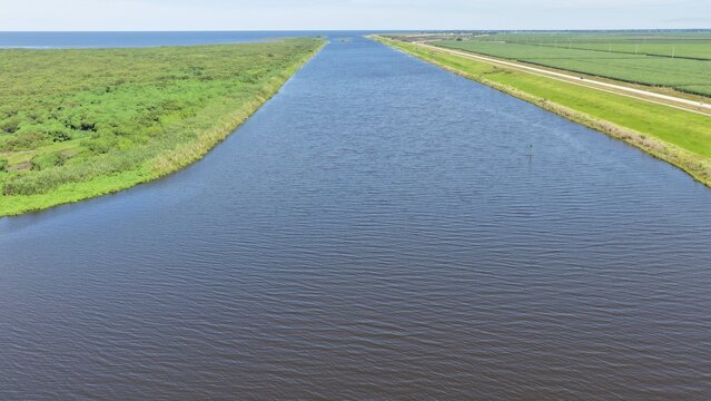 Aerial view of the canal near Lake Okeechobee with lush green vegetation and agricultural fields under a clear blue sky in Pahokee, Florida, United States.