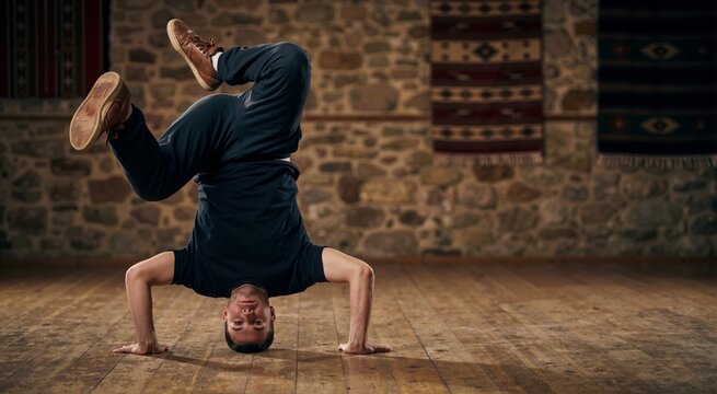 Breakdancer holding frozen headstand freeze pose on wooden floor. Athletic male bboy performing hip hop dance in studio