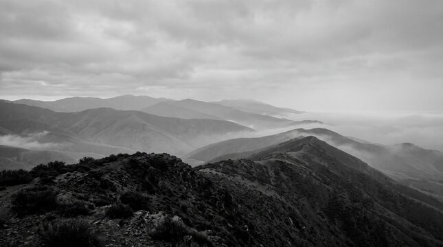 Black and white mountain range landscape with misty valleys and low clouds under overcast sky with distance beneath blanket layers mountain ridges fade distance
