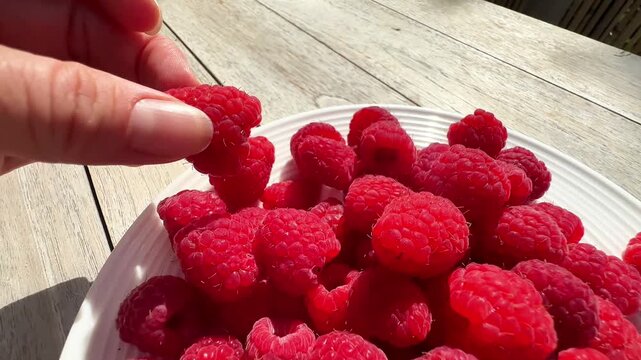  Hand picking a fresh raspberry from a white plate on a wooden table