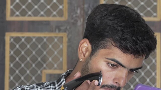 Young man trimming beard with an electric trimmer indoors