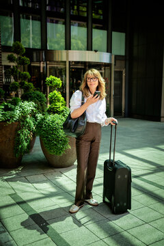 Confident mature businesswoman standing in front of the hotel entrance and using phone