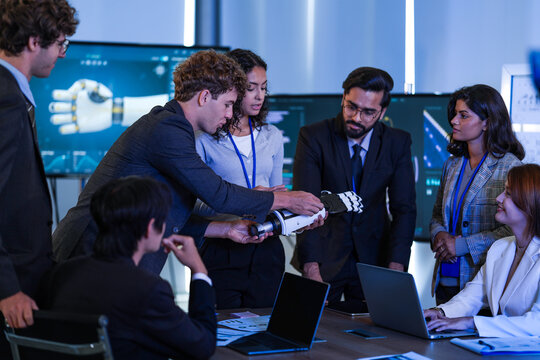 Seated bearded engineer in suit turns a bionic robotic arm while colleagues in suits and a woman in white blazer gather around to discuss it, a deep tech startup scene