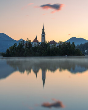 View of the Pilgrimage Church of the Assumption of Maria on Lake Bled with its reflection in the calm water at sunset, surrounded by mountains in Bled, Radovljica, Slovenia.