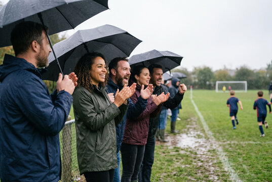 Parents with umbrellas cheering on the touchline of a kids' football game in the rain