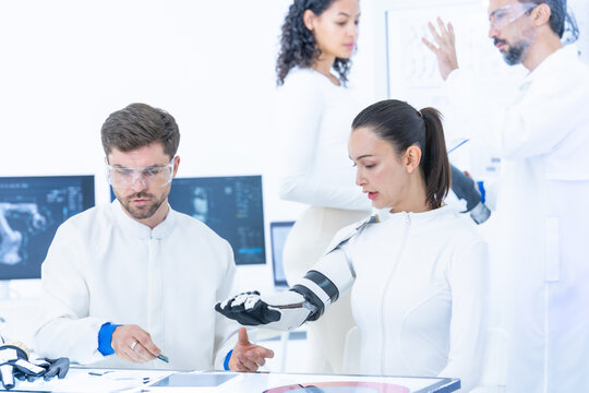 Robotics engineer seated at a lab bench fits a sensor-equipped exoskeleton forearm on a female test subject while aligning the forearm brace as a colleague records data on a tablet