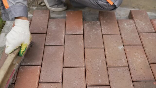 Close up video of a builder laying outdoor paving slabs. A worker in gloves uses a brush to sweep sand into the joints of red brick pavers to finish a new patio or walkway