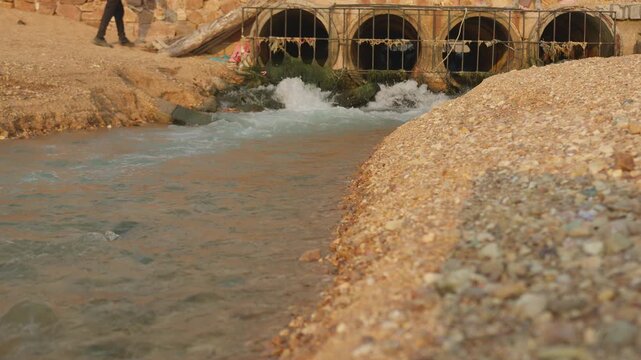 Wastewater runoff gushes from large concrete culvert pipes into a waterway, showcasing a storm drain system and its environmental impact on a gravel riverbank.