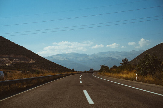 View of a highway stretching through a mountainous landscape with dry grass, power lines, and a clear blue sky in Greece.