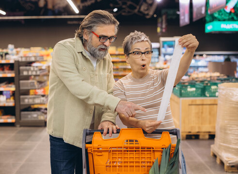 Senior couple looking at supermarket receipt, shocked by high grocery prices and inflation during shopping