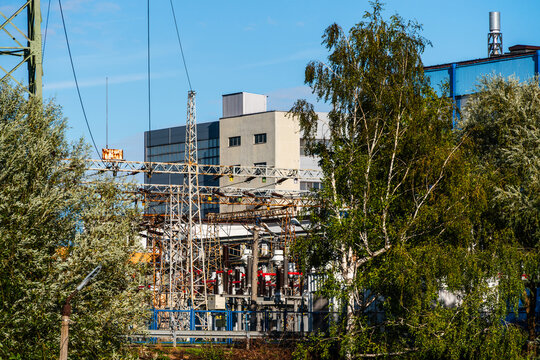 View of an electrical substation with high-voltage power lines and metal structures set against an industrial building and lush green trees in Poland.