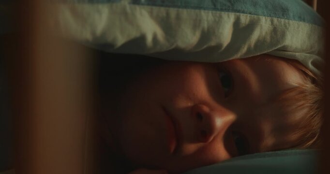 Eight-year-old boy hides completely under a soft blanket while lying in bed, then slowly peeks out and looks directly into the static camera in a warm and cozy bedroom. High quality 4k footage