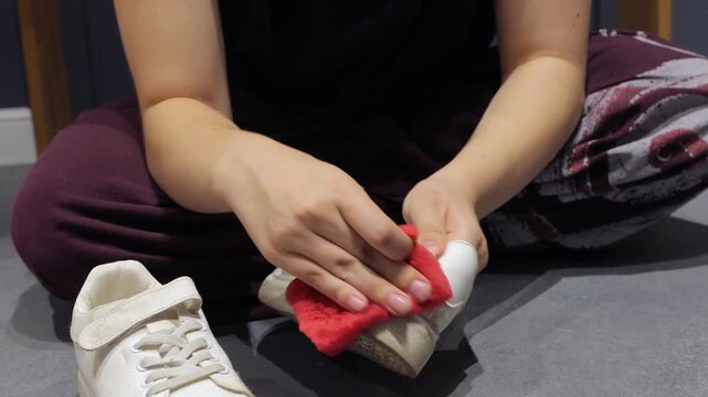 A person scrubs the side of a white sneaker with a red sponge on a wet tiled floor.