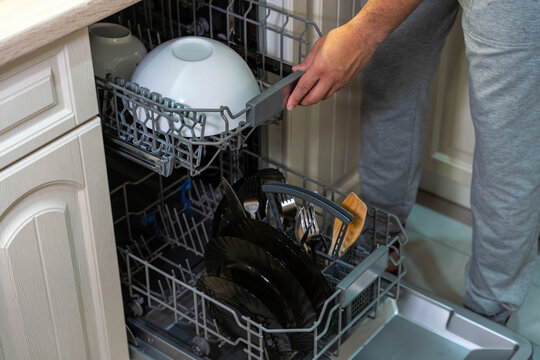 man organizing dishware inside household dishwasher machine, sliding open top basket holding ceramic bowl while lower section contains black dishes and cutlery in contemporary kitchen interior