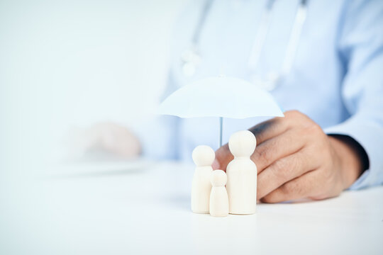 Doctor holding a white umbrella protecting wooden family figures on desk with copy space, concept of health insurance, family protection, medical care, and life security