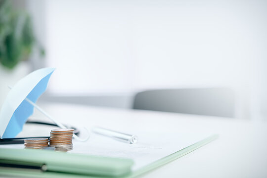 Stacks of coins protected by a small blue umbrella with financial documents on a white desk, concept of financial protection, insurance, wealth management, and risk savings.