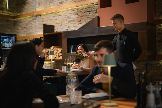 Waiter pouring wine for a couple toasting during a romantic anniversary dinner at a cozy luxury restaurant, side view.