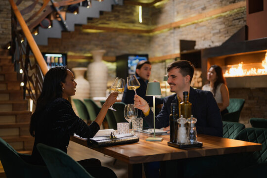 Interracial couple toasting, clinking wine glasses, bonding over fine dining at a busy luxury restaurant, side view.