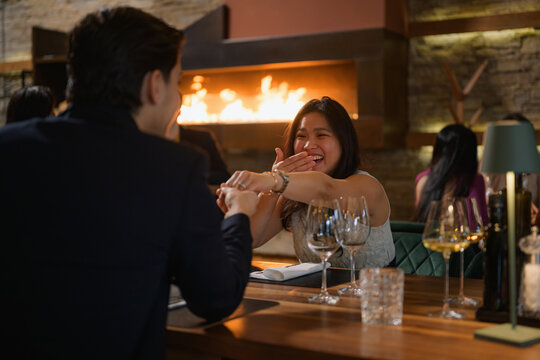 Emotional marriage proposal at a restaurant, a man giving an engagement ring to a smiling, surprised Asian woman.