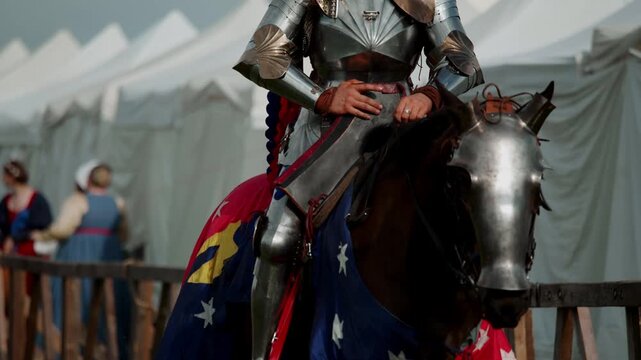 Armored knight rides decorated warhorse past white festival tents. Metal chamfron protects animal head while heavy cavalry soldier moves toward medieval jousting tournament arena.